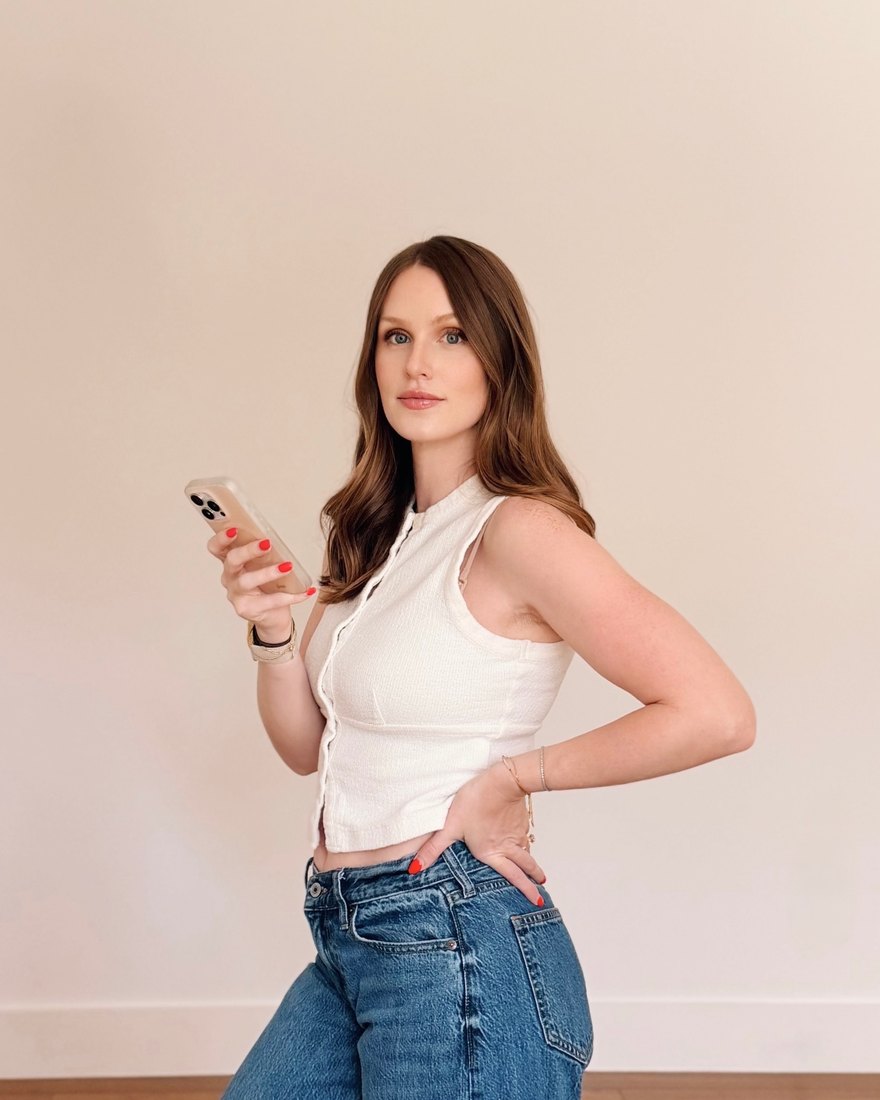 Rachel, a woman with long brown hair in a white sleeveless top and jeans, glancing over her shoulder at the camera with her phone in her hand